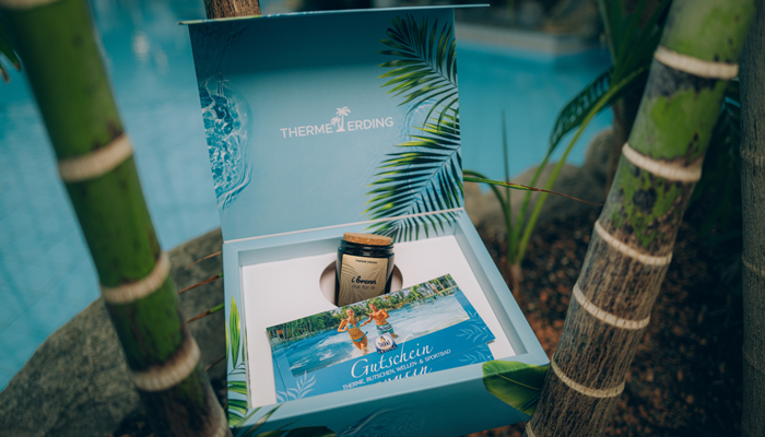 Geöffnete Geschenkbox der Therme Erding am Beckenrand mit Bambus im Hintergrund. In der Box liegen eine Duftkerze mit der Aufschrift „I brenn für di“ und ein Gutschein mit dem Schriftzug „Gutschein Therme Erding“. Auf der Innenseite des Deckels steht „THE