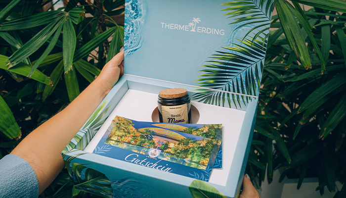 Geschenkbox mit blauem Hintergrund und Palmen, gefüllt mit zwei Gutscheinen und einer Kerze mit der Aufschrift "Beste Mama"