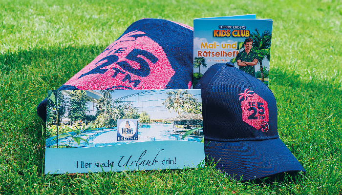 Geschenkumschlag, Cap mit pinkem Logo "TE 25 TM", Handtuch mit pinkem Logo "TE 25 TM" und Therme Erding Kids Club Mal- und Rätselheft liegen im Gras