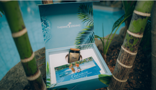 Geöffnete Geschenkbox der Therme Erding am Beckenrand mit Bambus im Hintergrund. In der Box liegen eine Duftkerze mit der Aufschrift „I brenn für di“ und ein Gutschein mit dem Schriftzug „Gutschein Therme Erding“. Auf der Innenseite des Deckels steht „THE